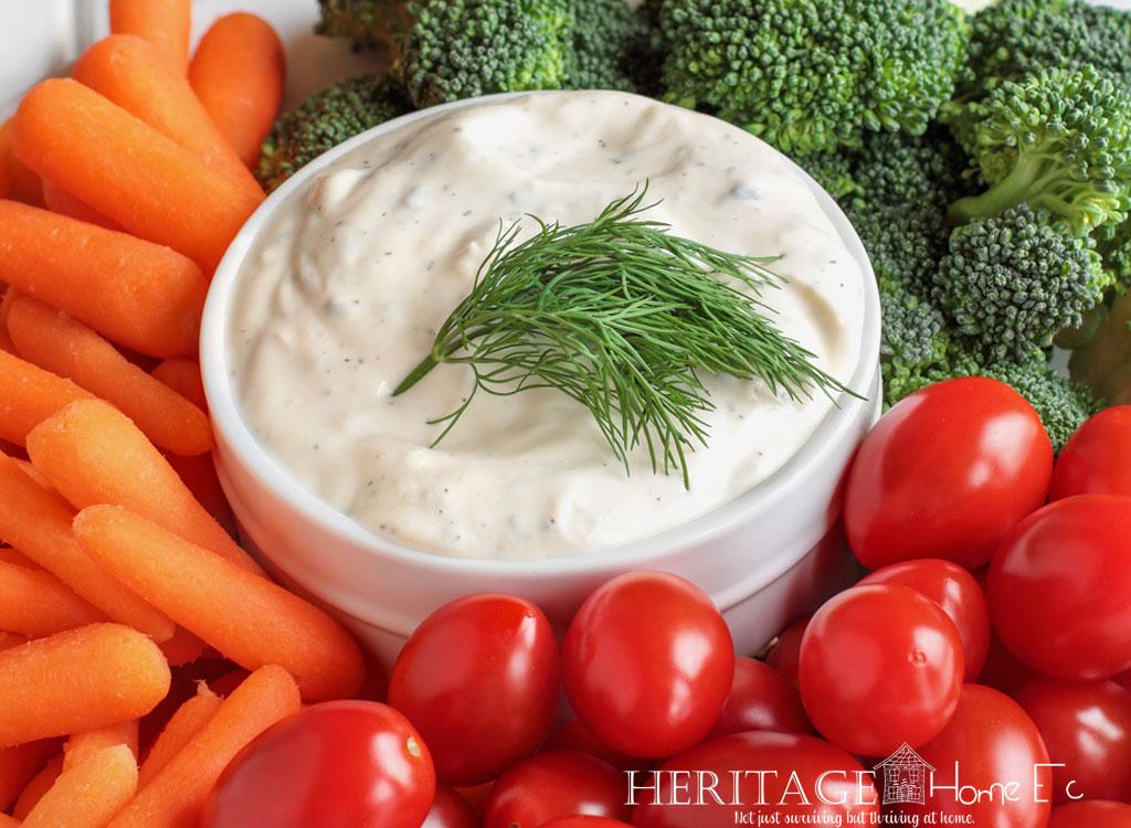 close-up of a white stoneware ramekin of Homemade Ranch Dressing in a ring of fresh baby carrots, cherry tomatoes, and broccoli garnished with a sprig of dill.